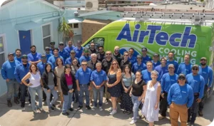A large group of people, some in blue AirTech uniforms, pose together in front of a green AirTech Cooling & Heating van outside a building.