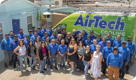 A large group of people, some in blue AirTech uniforms, pose together in front of a green AirTech Cooling & Heating van outside a building.