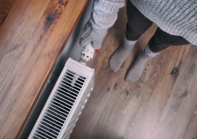 Person wearing gloves and socks adjusts the temperature knob on a white radiator heater, standing on a wooden floor near a wooden countertop.